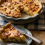 Quiche lorraine revisitée sans gluten dorée avec une part découpée révélant une garniture aux lardons et à la crème sur une planche en bois et une serviette a carreau