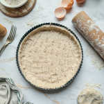 Fond de tarte en pâte brisée au sarrasin sans gluten piqué à la fourchette dans un moule noir, entouré d'un rouleau à pâtisserie et de coquilles d'œufs.