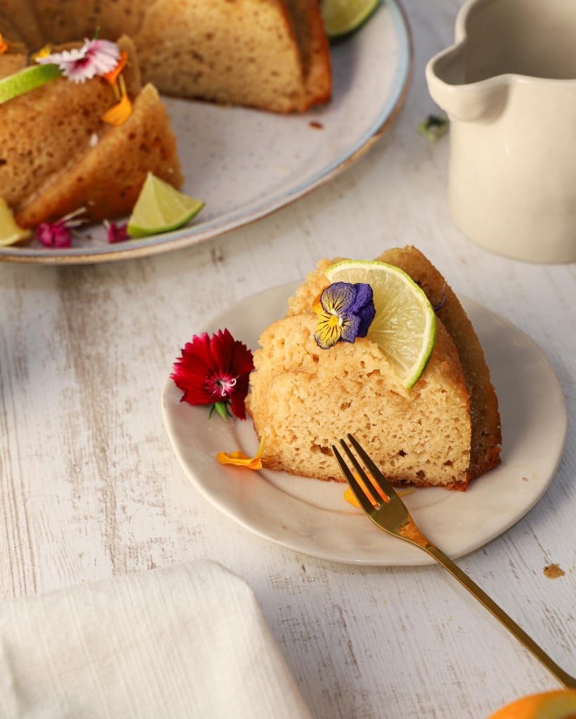 Une part de moelleux au citron sans gluten et sans lactose décorée d'une tranche de citron vert et de fleurs comestibles, servie sur une petite assiette blanche avec une fourchette dorée