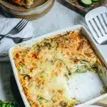 Gratin de courgettes sans gluten doré et gratiné dans un plat blanc avec spatule Ingrédients pour gratin de courgettes sans gluten : courgettes, crème de soja, oeufs, fromage râpé