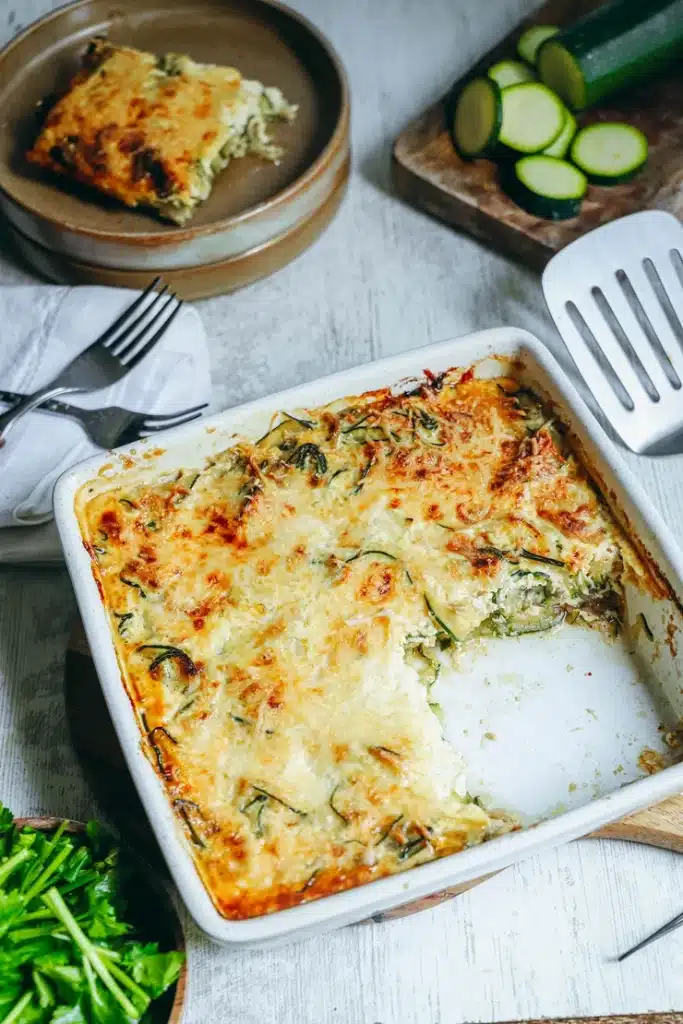 Gratin de courgettes sans gluten doré et gratiné dans un plat blanc avec spatule Ingrédients pour gratin de courgettes sans gluten : courgettes, crème de soja, oeufs, fromage râpé