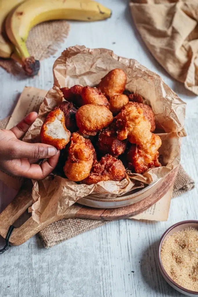 Beignets à la banane sans gluten sans lactose dorés et croustillants dans un bol en papier kraft avec main qui en prend un