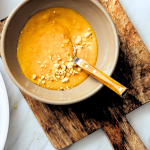Sauce crémeuse à la cacahuète sans gluten dans un bol beige avec cacahuètes concassées et cuillère en bois sur planche en bois