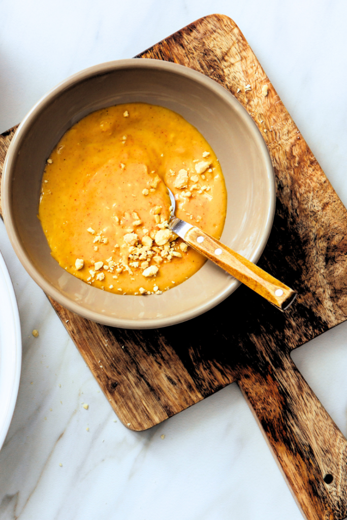Sauce crémeuse à la cacahuète sans gluten dans un bol beige avec cacahuètes concassées et cuillère en bois sur planche en bois